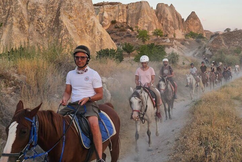 A group of people riding horses on a dusty trail with rocky formations in the background. 3