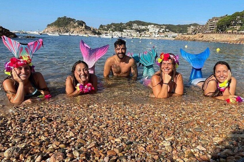 Personas en aguas poco profundas con coloridas colas de sirena, playa de guijarros y un pueblo costero al fondo. 4