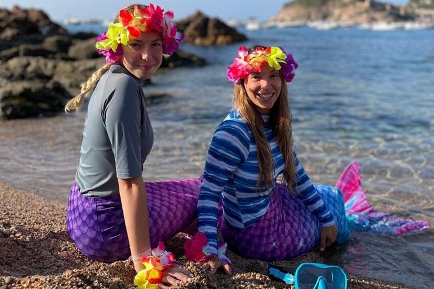 Dos personas sentadas en una playa con costa rocosa y el océano al fondo. 1