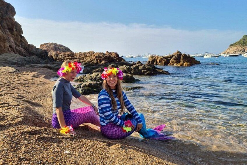 Dos personas sentadas en una playa rocosa con aguas cristalinas y barcos al fondo. 3
