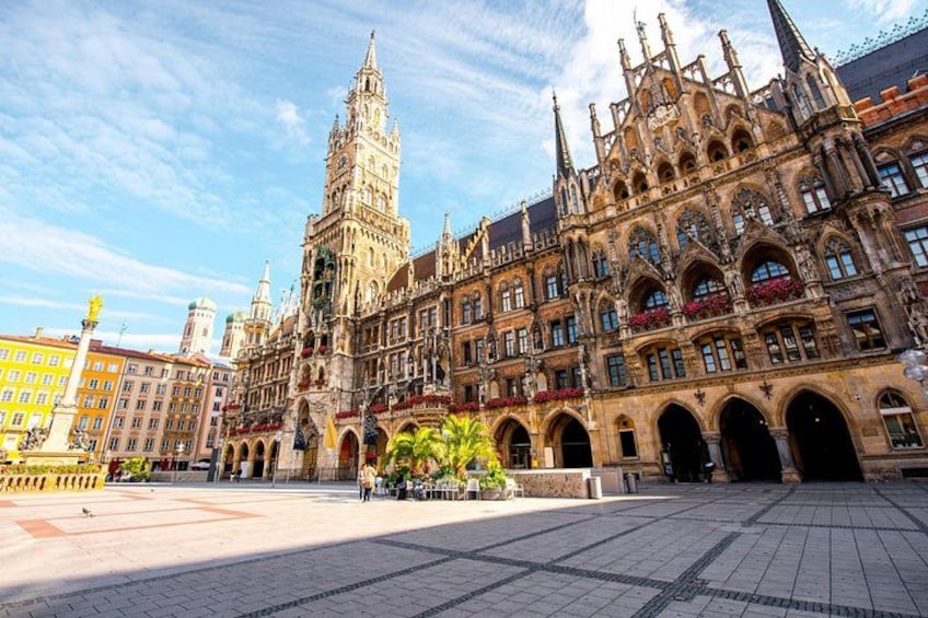 A historic building with intricate architecture, a prominent tower, and a paved square with a fountain. 3