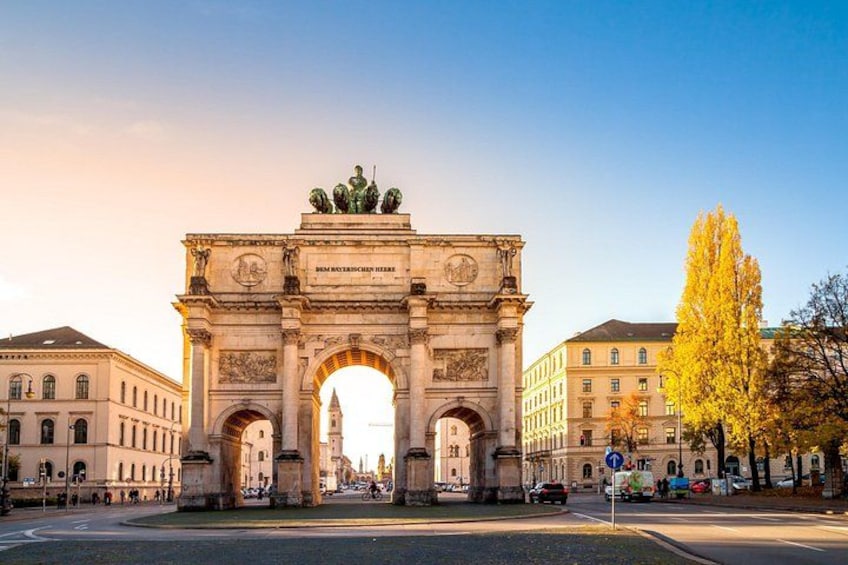 A historic archway with inscriptions, flanked by classical buildings, under a clear sky. 1