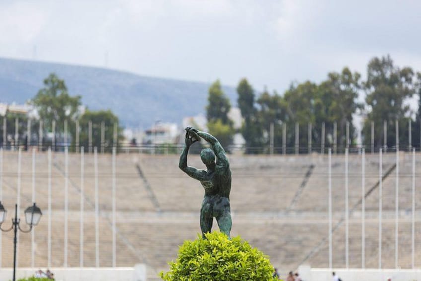 A statue of a person holding a shield, set on a pedestal with a fence and trees in the background. 2