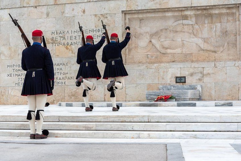 Guards in uniform standing in formation, a stone wall with inscriptions, and a statue in the background. 4