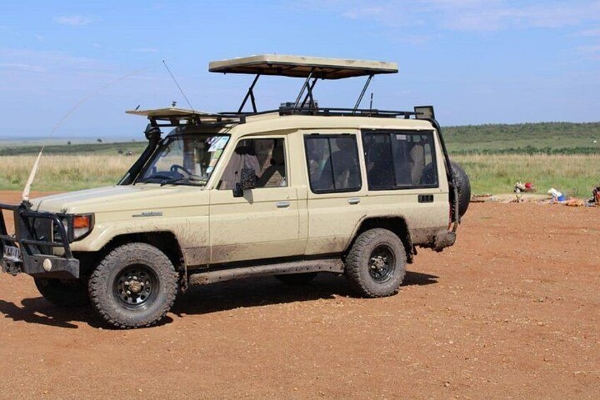 A beige off-road vehicle with a roof rack, parked on a dirt road with a grassy field and hills in the background. 4