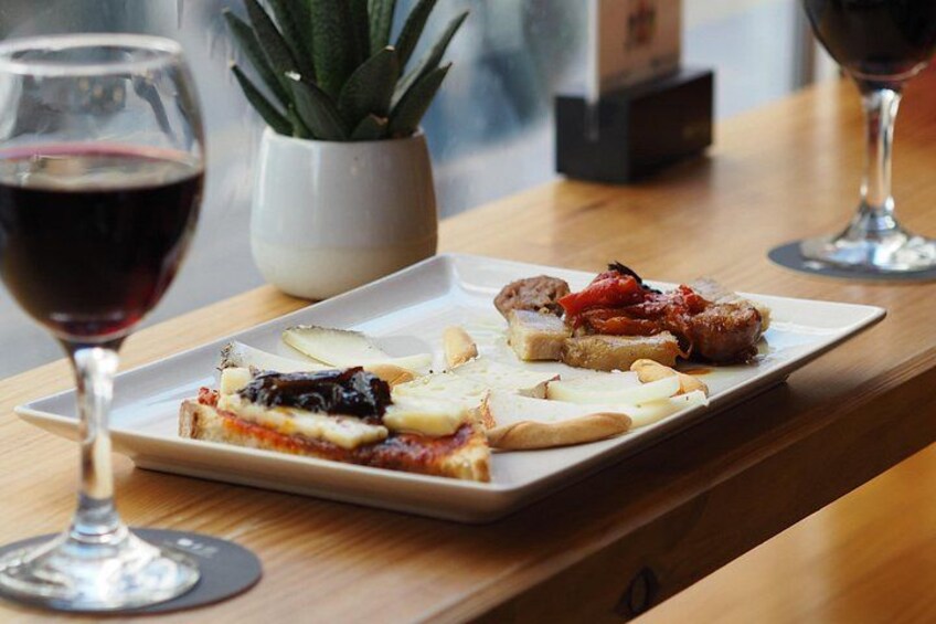 A plate of food with a slice of pizza, a piece of bread, and a small dish of sauce, accompanied by a glass of red wine on a wooden table. 2