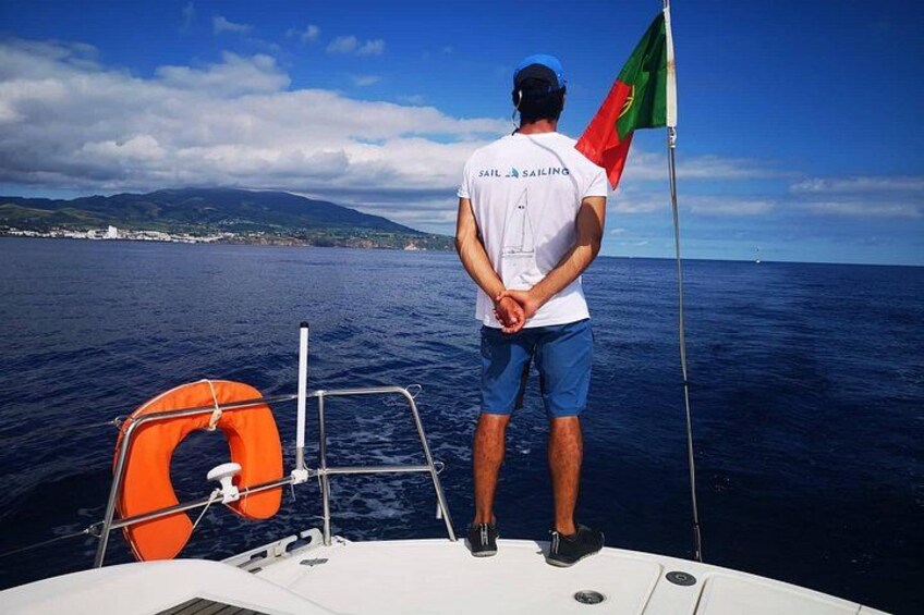 A person on a boat wearing a white shirt with 'Sail Sailing' text, standing on the deck with a flag. 4