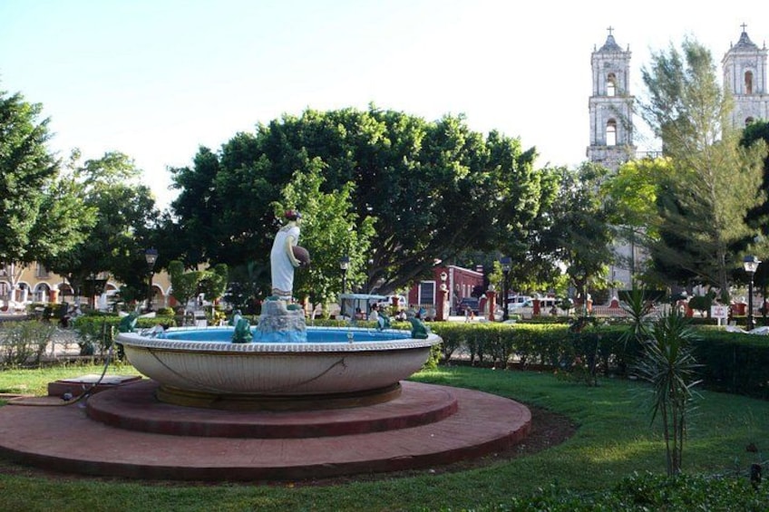 Une fontaine ornée d’une statue au centre, entourée d’une plateforme circulaire, avec une église et des arbres en arrière-plan. 4