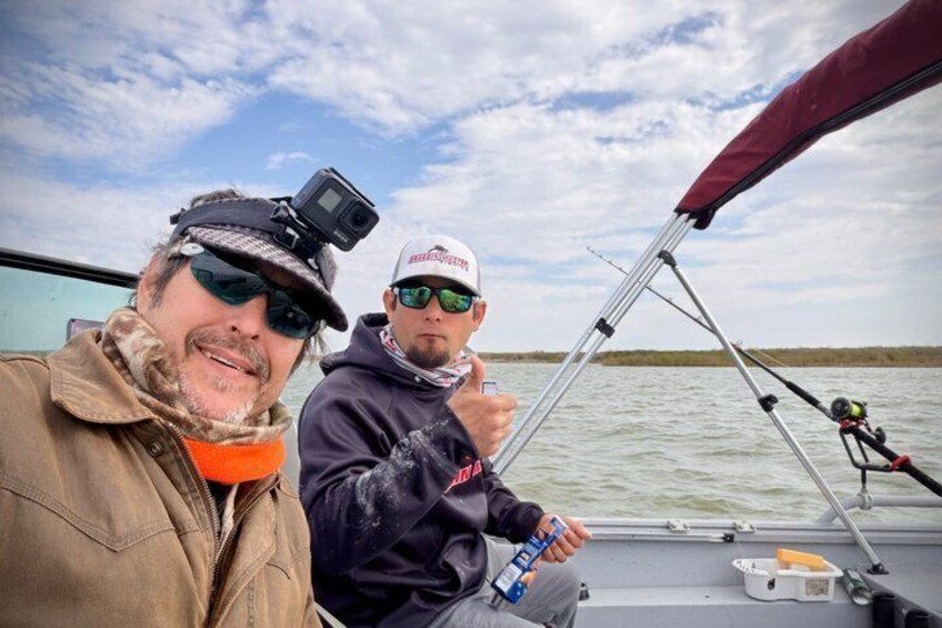 Two people on a boat with fishing rods, a camera, and a water body in the background. 5