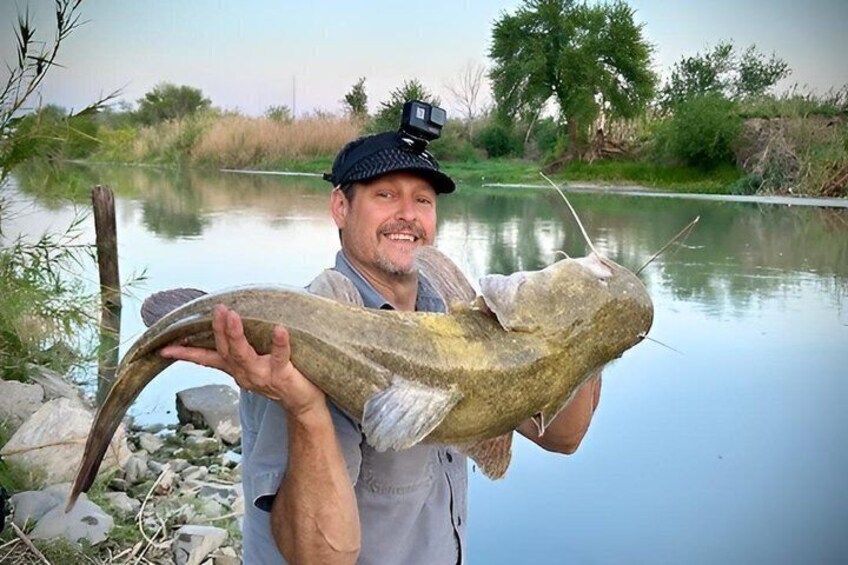 A person holding a large fish by a calm river. 4