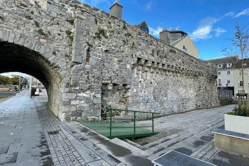 A stone bridge with a green railing, a paved walkway, and buildings in the background. 1