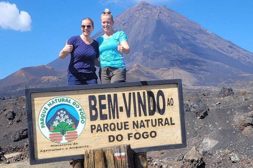 Dos personas frente a un letrero que dice 'Bem-Vindo ao Parque Natural do Fogo'. 1