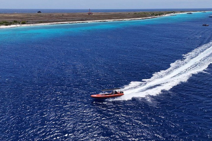 A boat speeding across a deep blue sea with a wake, surrounded by a coastline with sandy beaches and shallow waters. 3