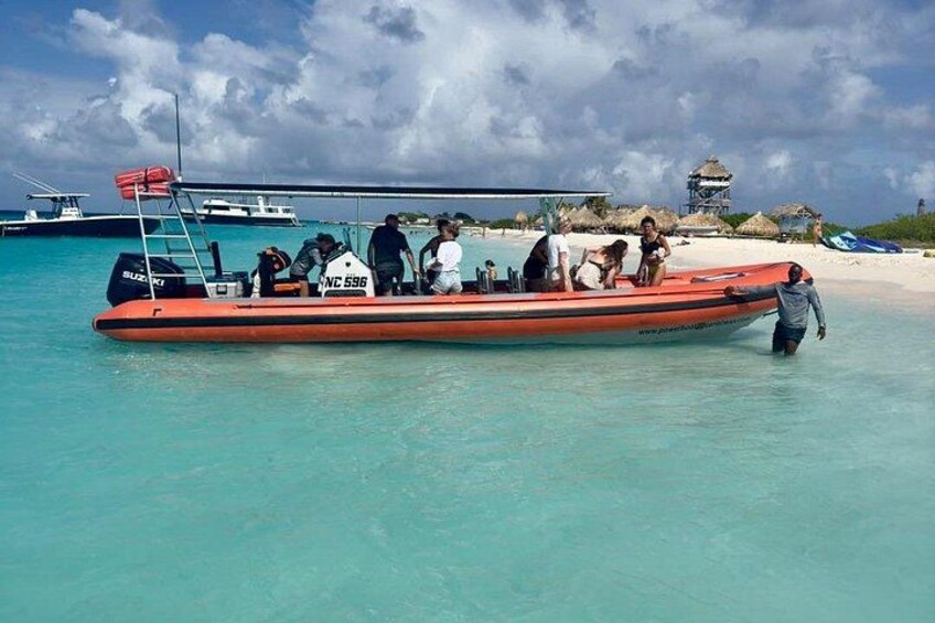 A red boat with people on a clear turquoise sea, a sandy beach, and a thatched-roof structure in the background. 5