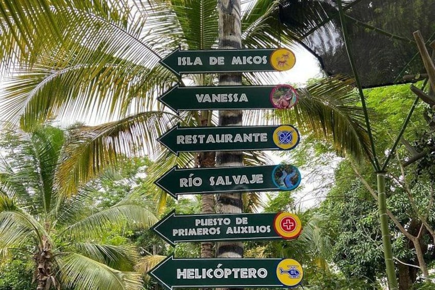 A directional signpost with multiple arrows pointing to various destinations such as Isla De Micos, Vannesha, Restaurante, Rio Salvaje, Primero De Auxilios, and Helicoptero. 5