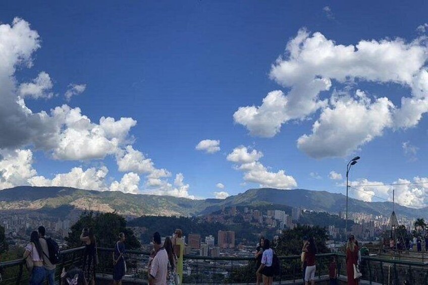 Eine Gruppe von Menschen auf einer Brücke mit Blick auf eine Stadtlandschaft und dahinter Bergen. 3