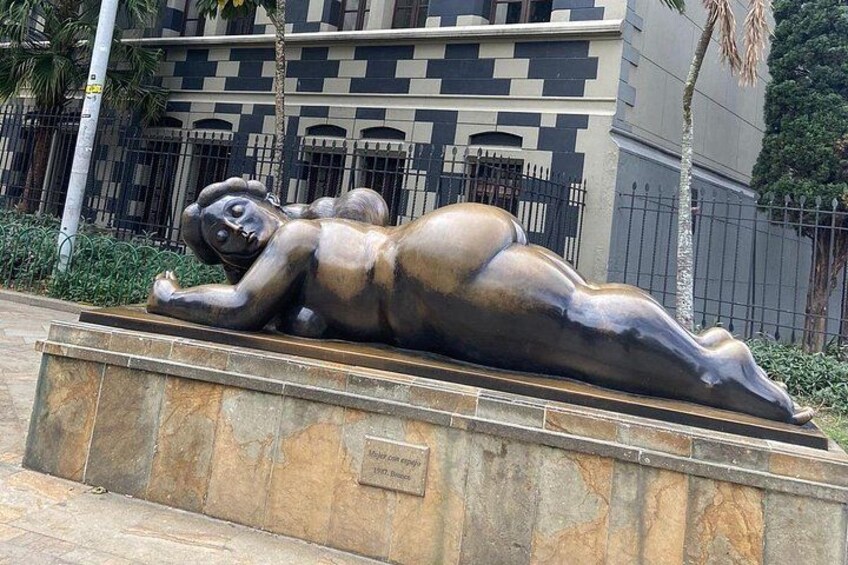 Eine bronzene Skulptur einer liegenden Figur auf einem Sockel, im Hintergrund ein Gebäude. 1
