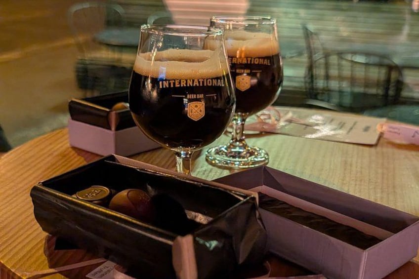 Two glasses of dark beer with the label 'International Beer Bar' on a table with a box of chocolates and a menu. 2