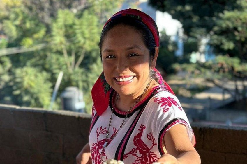 A person wearing a traditional embroidered blouse with red patterns, standing outdoors with trees and a building in the background. 1
