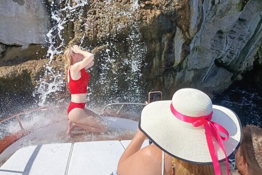 A woman in a red swimsuit on a boat, splashing water, with a rocky cliff in the background. 3