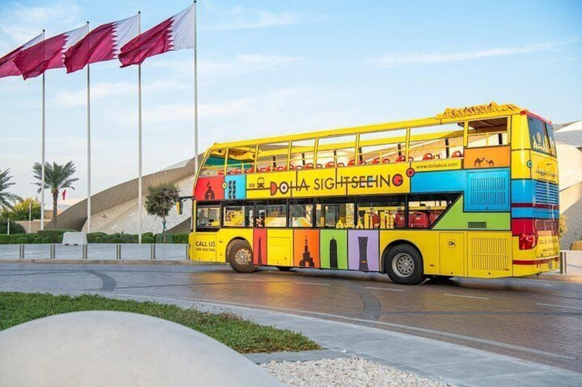 A double-decker bus with colorful designs and the text 'Doha Sightseeing' parked in front of a modern building with flags. 5