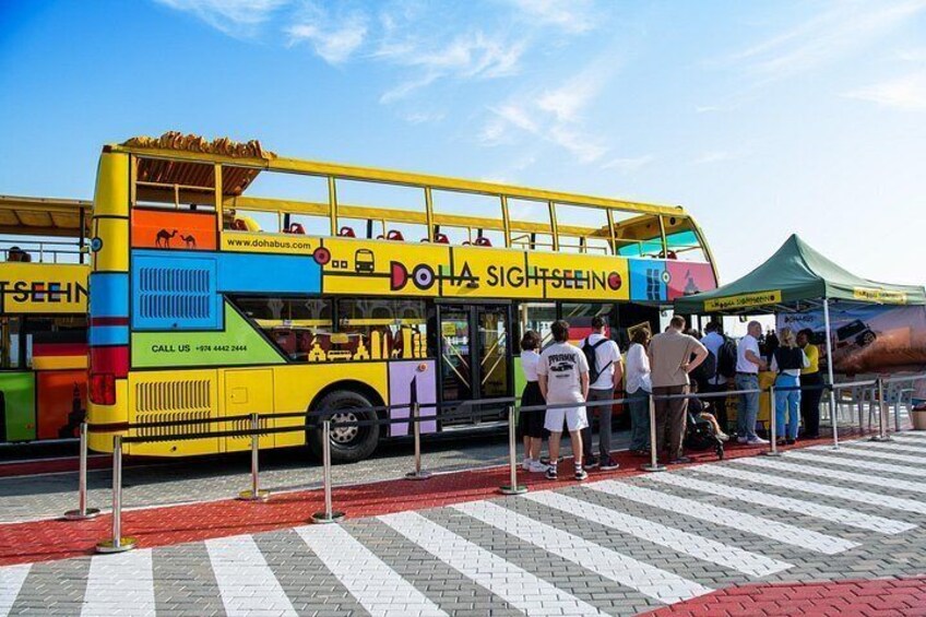 A colorful double-decker bus with 'DOHA SIGHTSEEING' written on it, parked with people waiting in line. 2