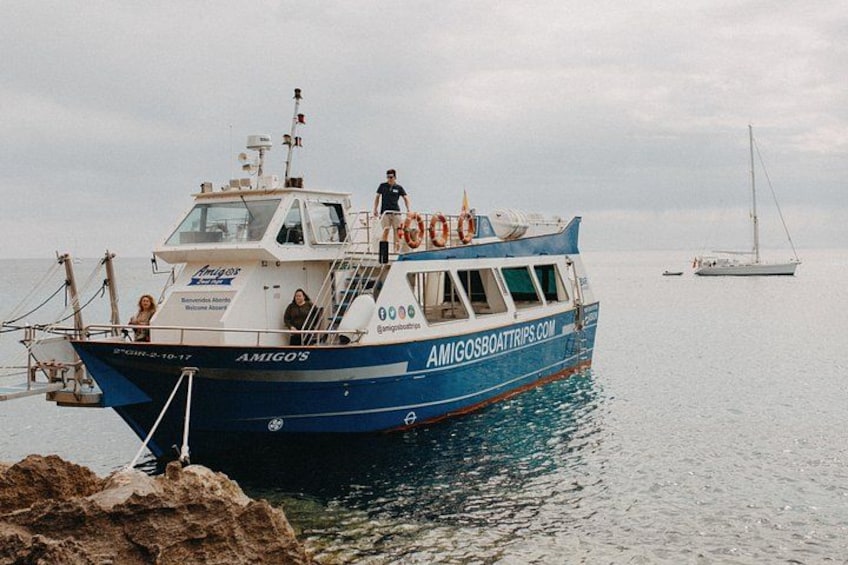 Un bateau bleu et blanc, baptisé AMIGO'S, était amarré sur une côte rocheuse, avec un voilier visible au loin. 4