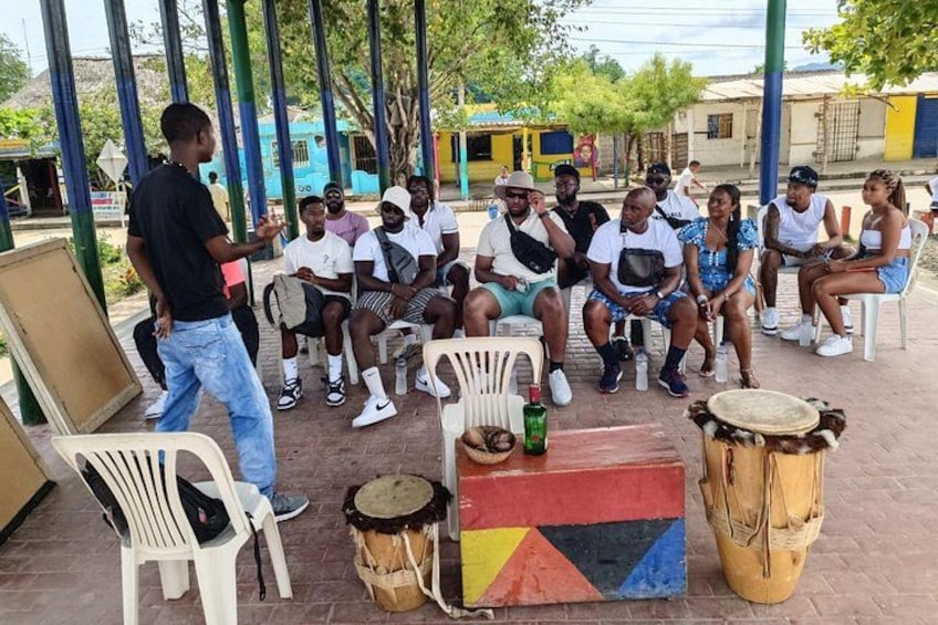 A group of people sitting on plastic chairs outdoors, with drums and a colorful block in the foreground. 1