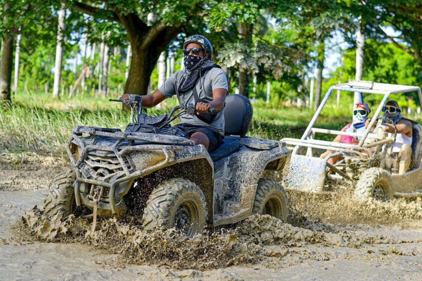 A person riding an ATV through a muddy area with another ATV in the background. 1