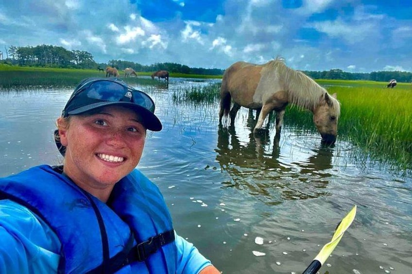 A person in a blue jacket and cap taking a photo with a horse in a body of water. 2