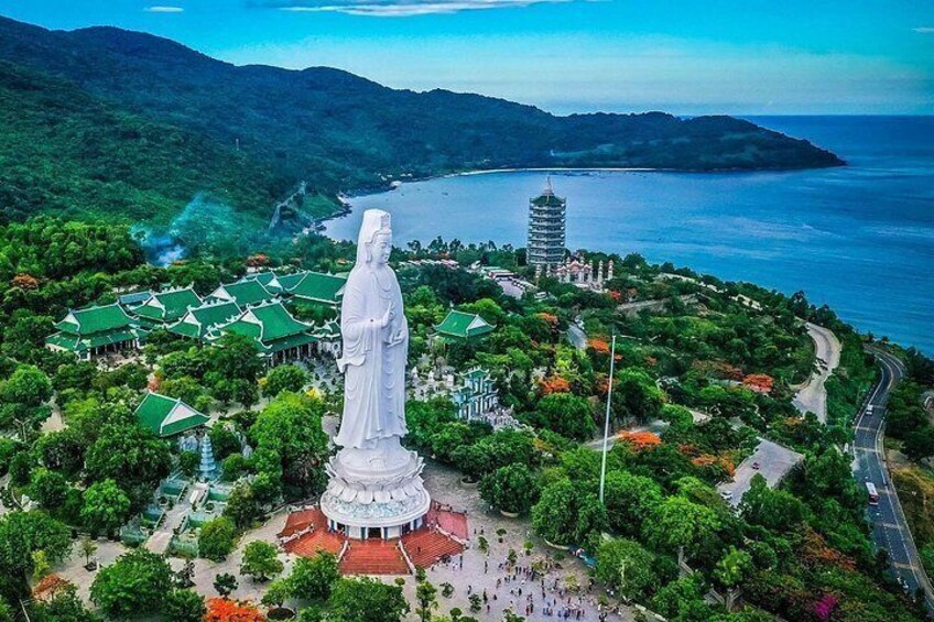 A large white statue surrounded by lush greenery and traditional architecture, with a coastal view in the background. 1