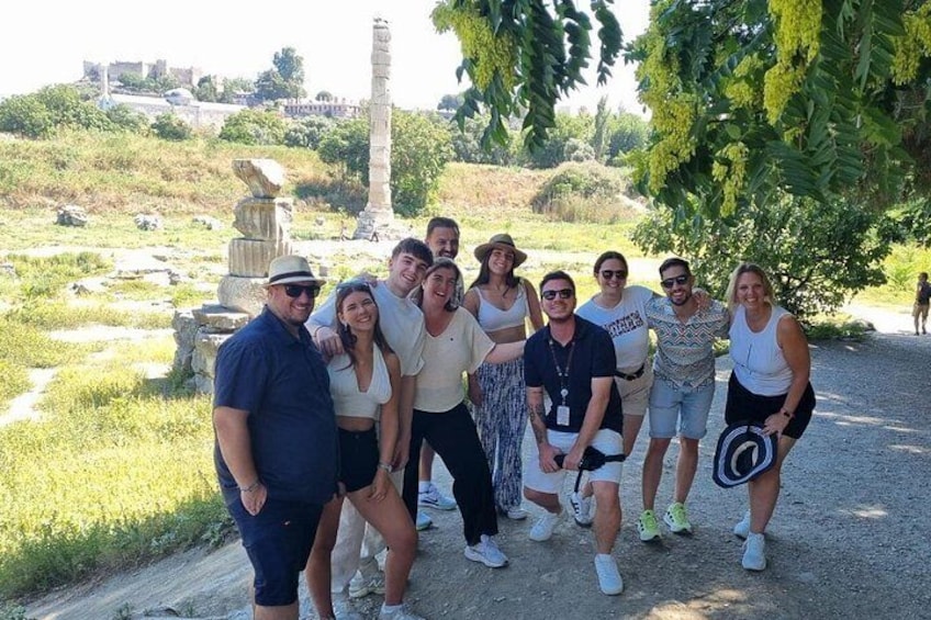 A group of people standing on a dirt path with ancient ruins and greenery in the background. 1