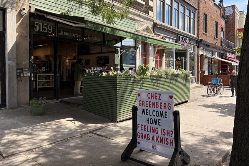 En butik med ett skyltfönster där det står ”Chez Greenberg, välkommen hem” och en vägskylt med texten ”Känner du dig lite nere? Köp en knish.” 2