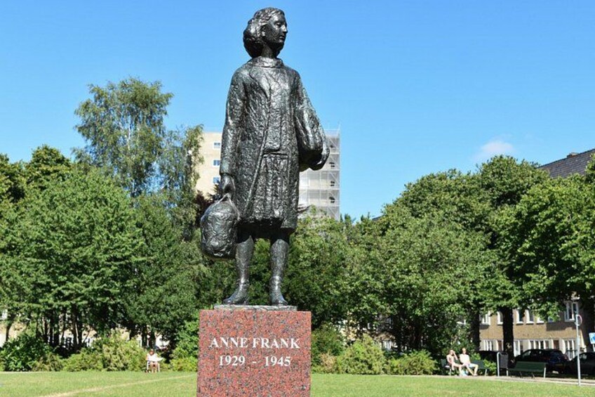 A statue of Anne Frank holding a suitcase, with a plaque reading 'Anne Frank 1929-1945' in a park with trees and buildings in the background. 2