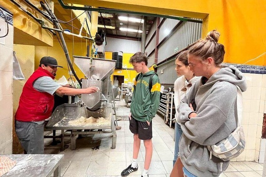 Un área de procesamiento de alimentos con un trabajador operando una máquina en forma de embudo grande, una persona joven con una chaqueta verde observando, y dos personas de pie cerca. 4