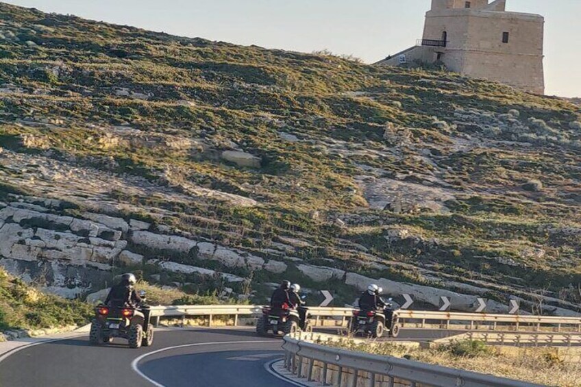 Una carretera sinuosa con motociclistas, una barandilla y una torre en una colina. 2
