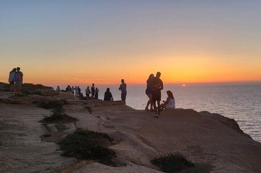 Personas de pie y sentadas en una costa rocosa al atardecer. 2