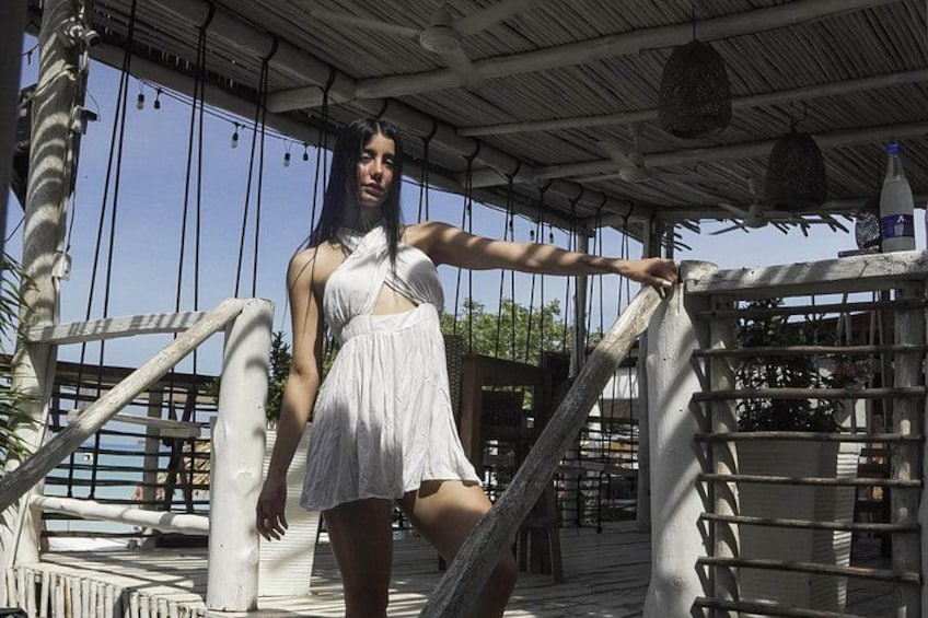 A person in a white dress standing on a wooden deck with a metal railing, under a covered area with hanging lights. 2