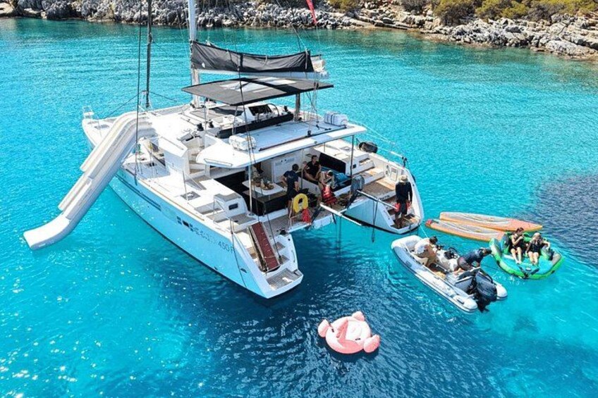 A white catamaran with a slide, people on board, and a pink inflatable on the water. 1