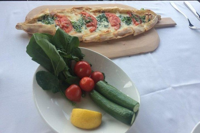 A long, rectangular flatbread with tomato and spinach toppings, a plate with cherry tomatoes, cucumber slices, and a lemon wedge, and a wooden cutting board. 3