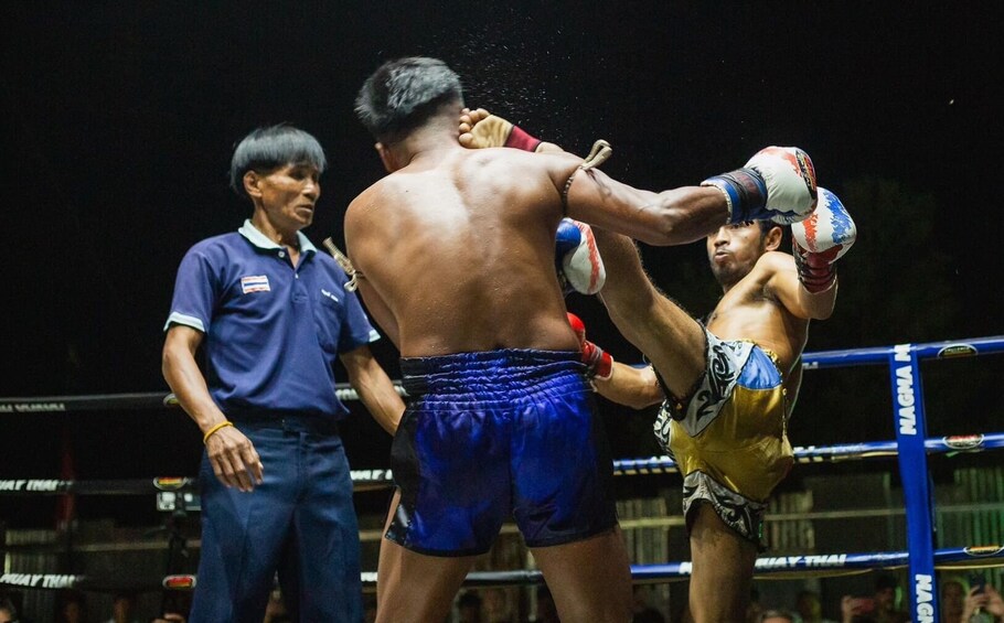 Combate de Muay Thai con dos luchadores en un ring, uno con shorts azules y el otro con shorts amarillos, y un árbitro con camisa azul. 3