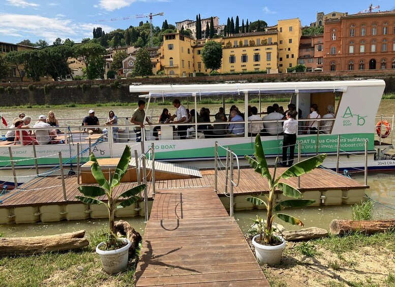 Una embarcación con pasajeros atracada en un muelle de madera con plantas en macetas, con edificios y un cielo despejado al fondo. 2