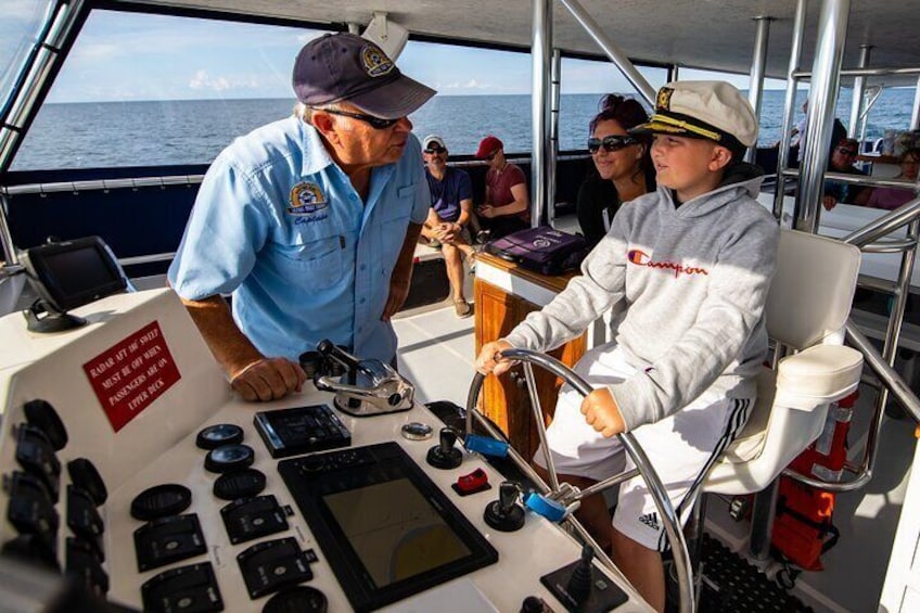 A boat's control station with a person steering and another person seated, a steering wheel, and various controls. 4