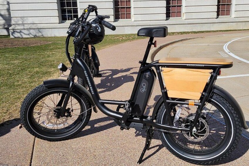 A black electric bicycle with a wooden seat and a rear rack, parked on a paved surface. 5
