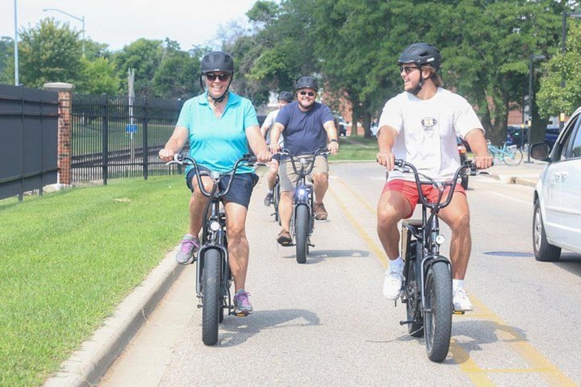 Three cyclists on a paved path with grass on the sides, a fence, and trees in the background. 3