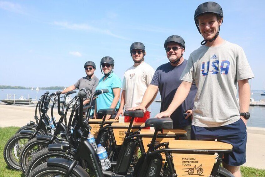 A group of people standing by a row of bicycles, with a body of water and boats in the background. 5