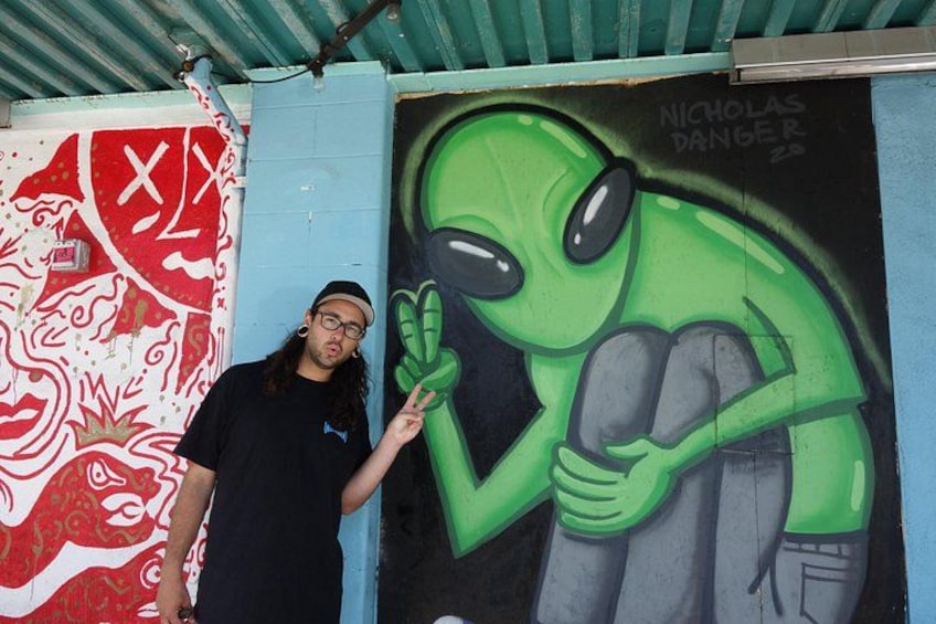 Una persona de pie junto a un mural de un extraterrestre verde haciendo un gesto con la mano. 2