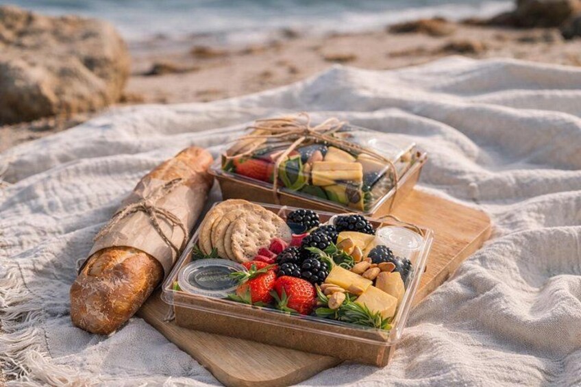 A picnic setup on a beach with a baguette, a container of fruits and nuts, and a wooden board. 2
