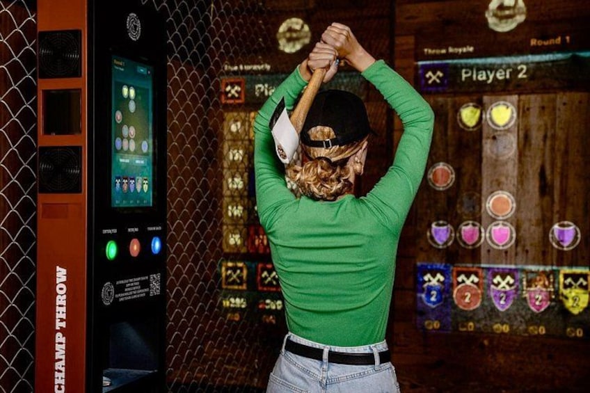 A person in a green shirt and cap holding a bat, standing in front of a bowling lane with a scoreboard and a chain-link fence. 5