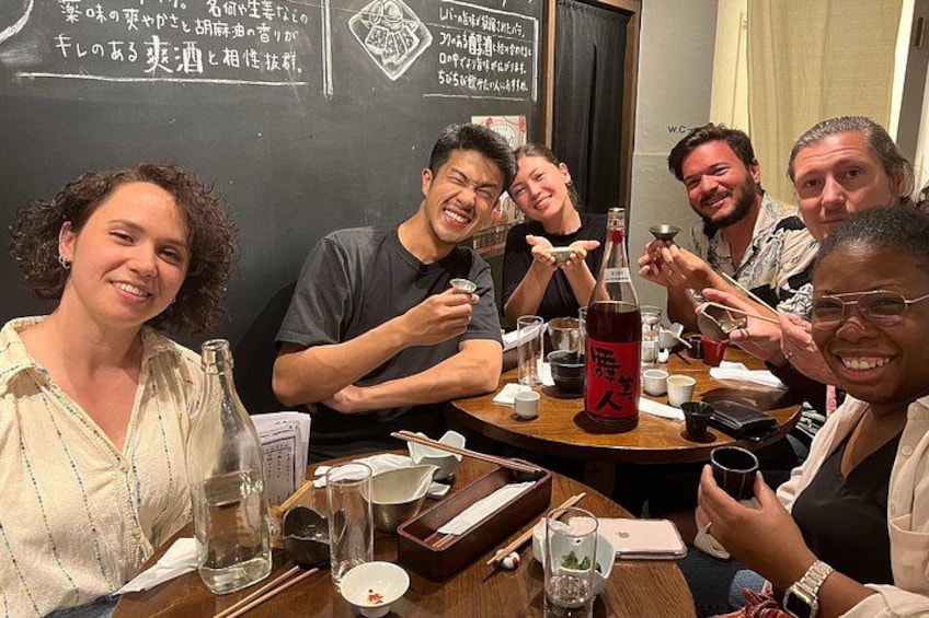 A group of people sitting around a table with various dishes and drinks, including a bottle of red beverage. 3